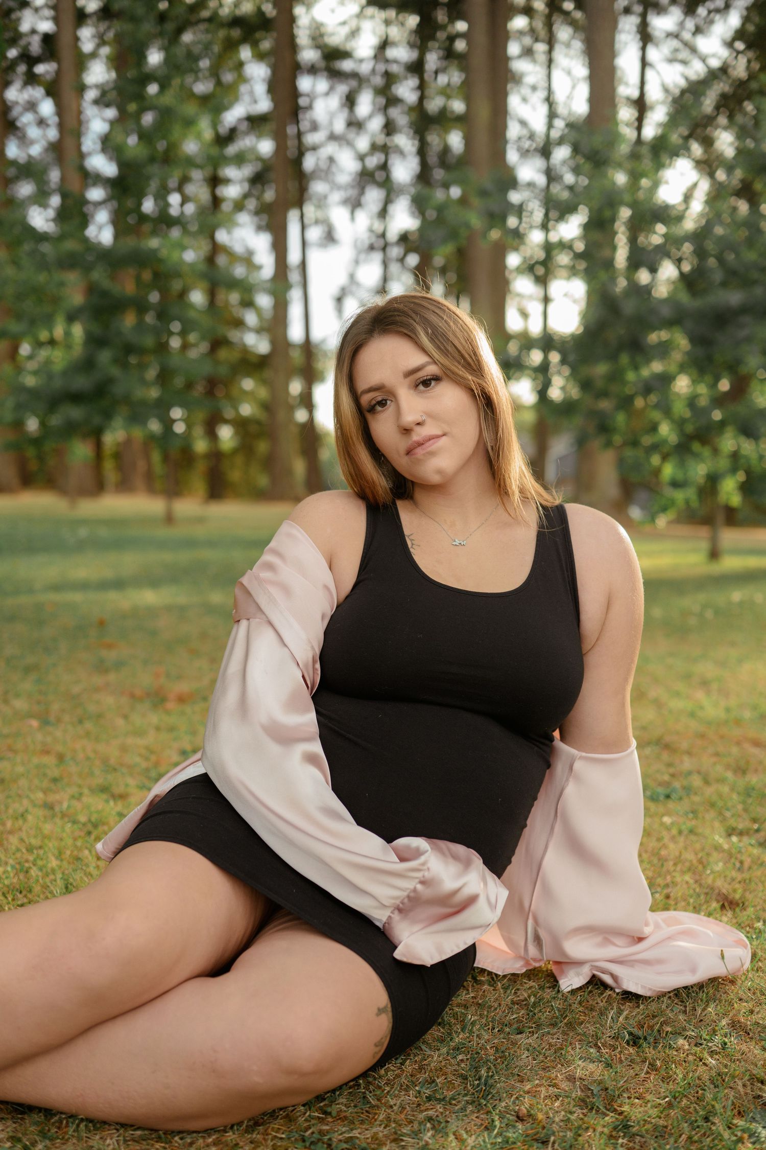 Someone in a black dress and pink cardigan poses on grass in a sunny wooded park setting.