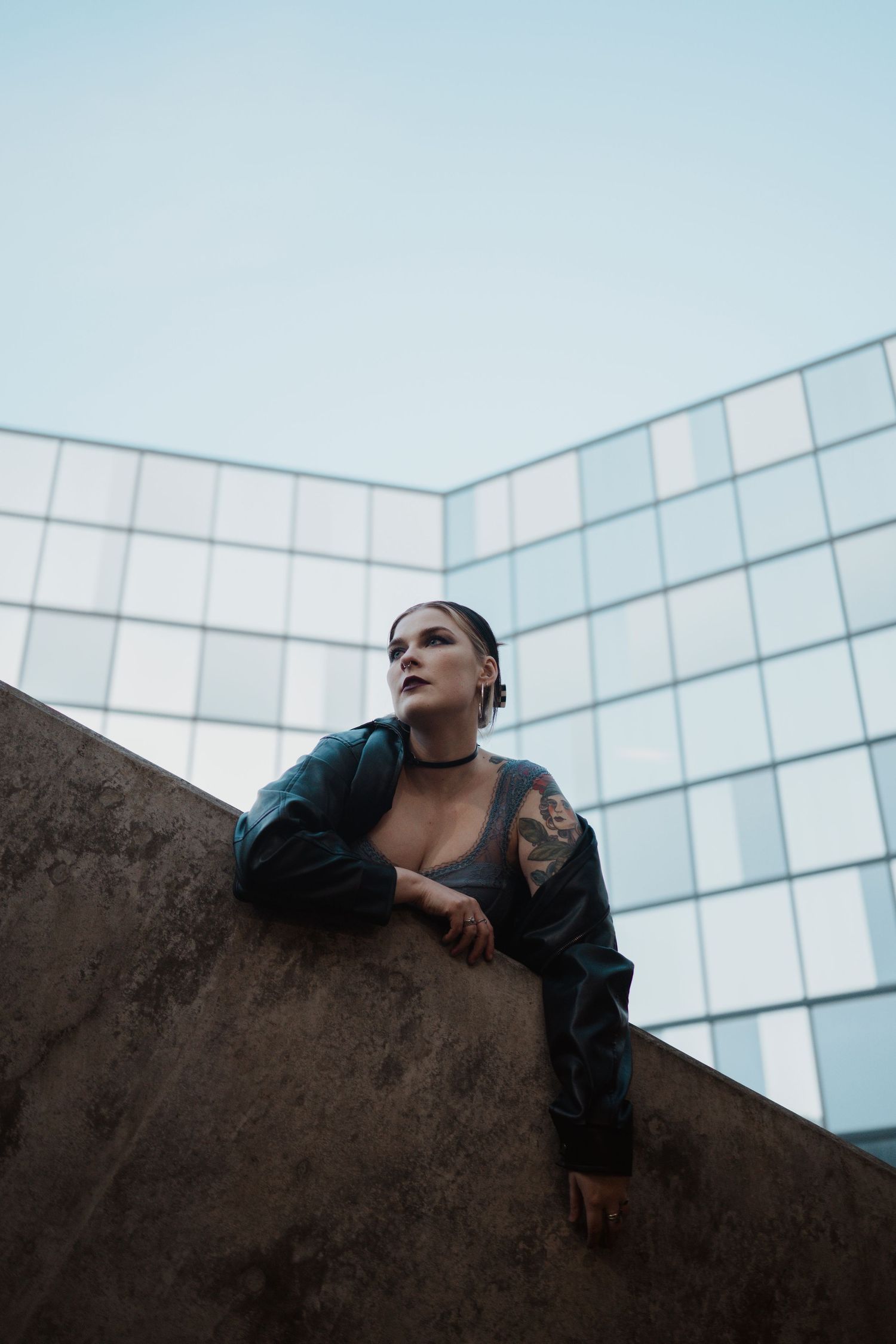 Figure in dark outfit poses against modern glass building architecture at dusk.