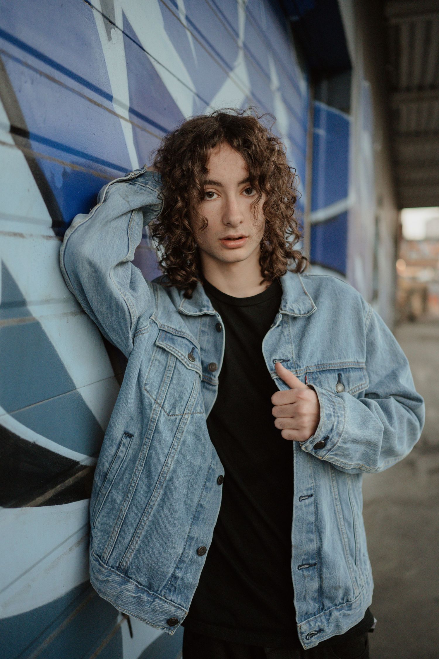 A casual street style photo featuring denim jacket and curly hair against an urban wall with blue artwork.