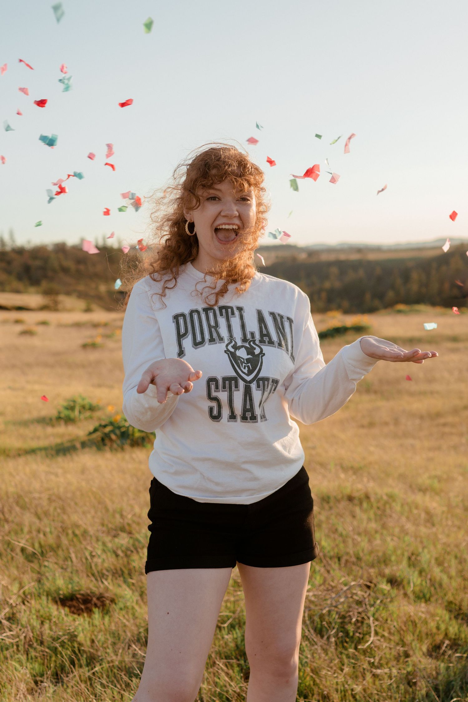 Person in Portland State sweatshirt throws confetti in sunny field.