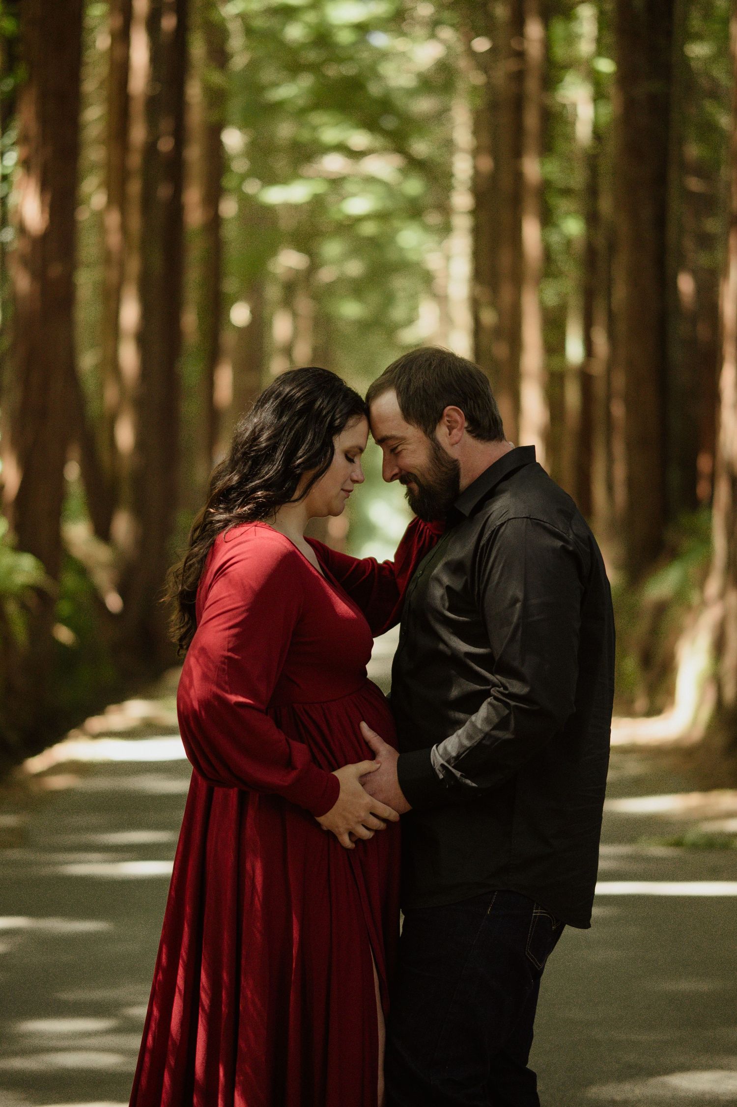 A romantic couple in a forest path shares an intimate moment as they await their baby, wearing red and black outfits.