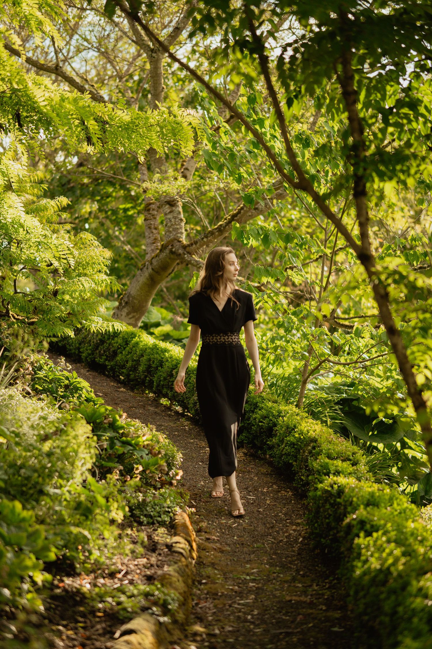 A person in a black dress walks along a winding garden path surrounded by lush green foliage on a sunny day.