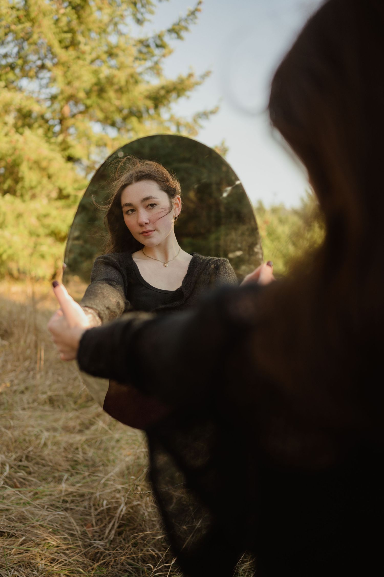 Creative portrait reflected in circular mirror held outdoors with evergreen trees in background.