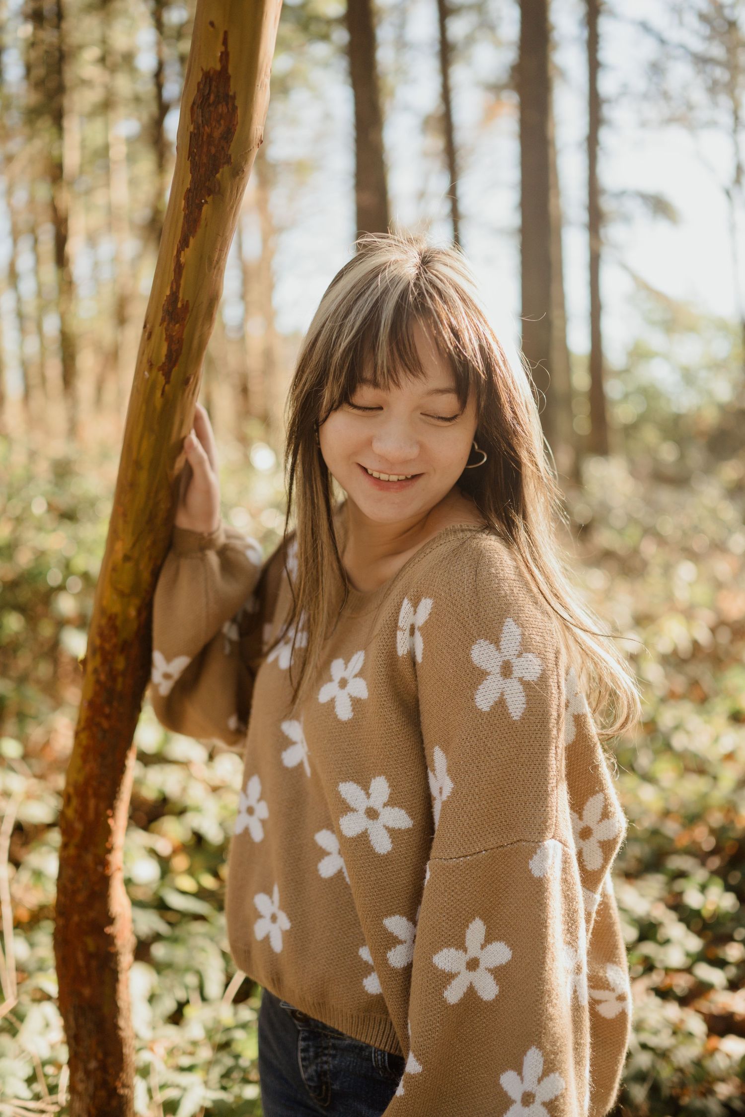 Person in tan sweater with white flower pattern smiling in sunlit forest.