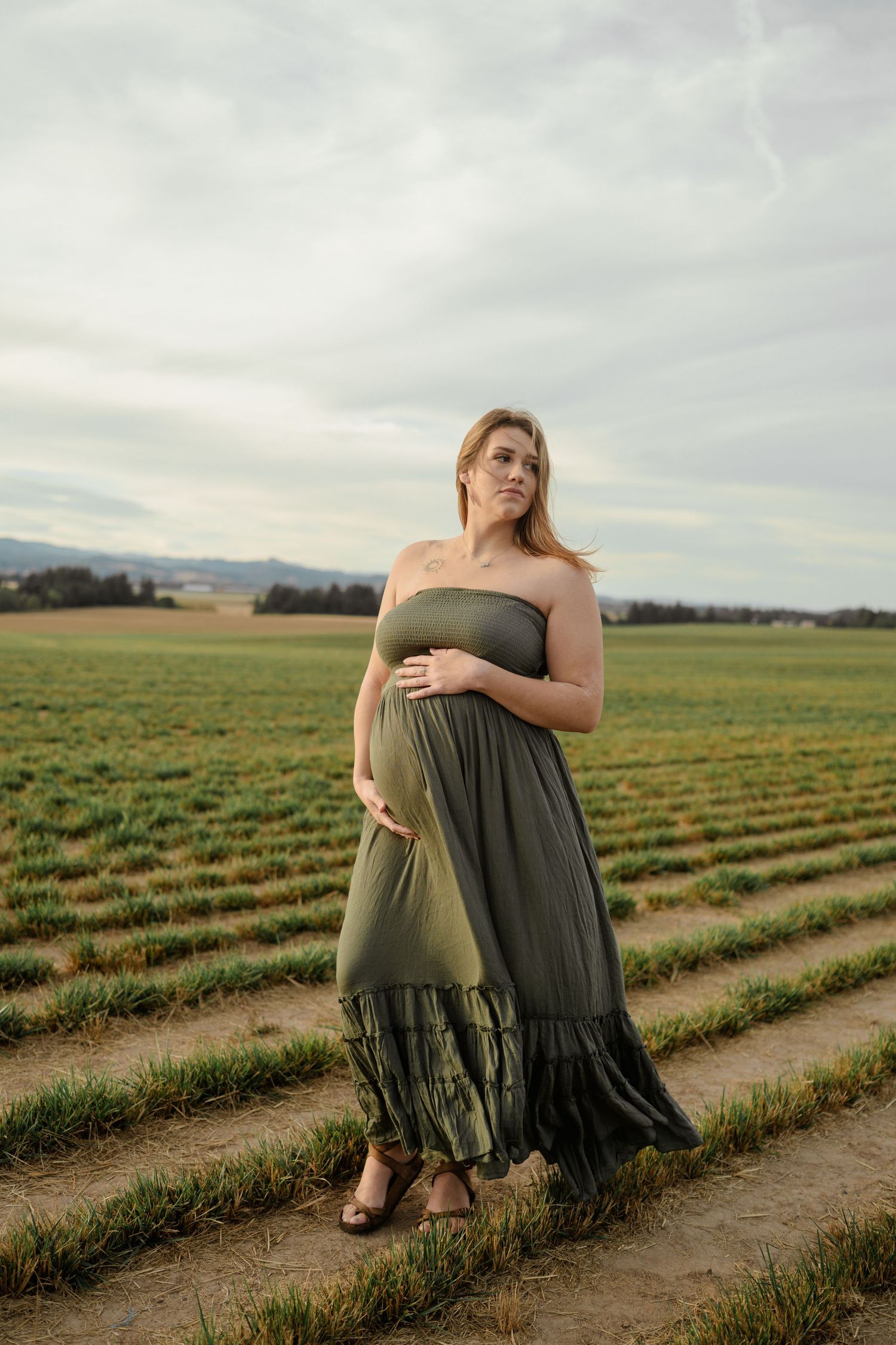 A woman in an olive maxi dress stands in a scenic agricultural field with mountains in the distance during golden hour.