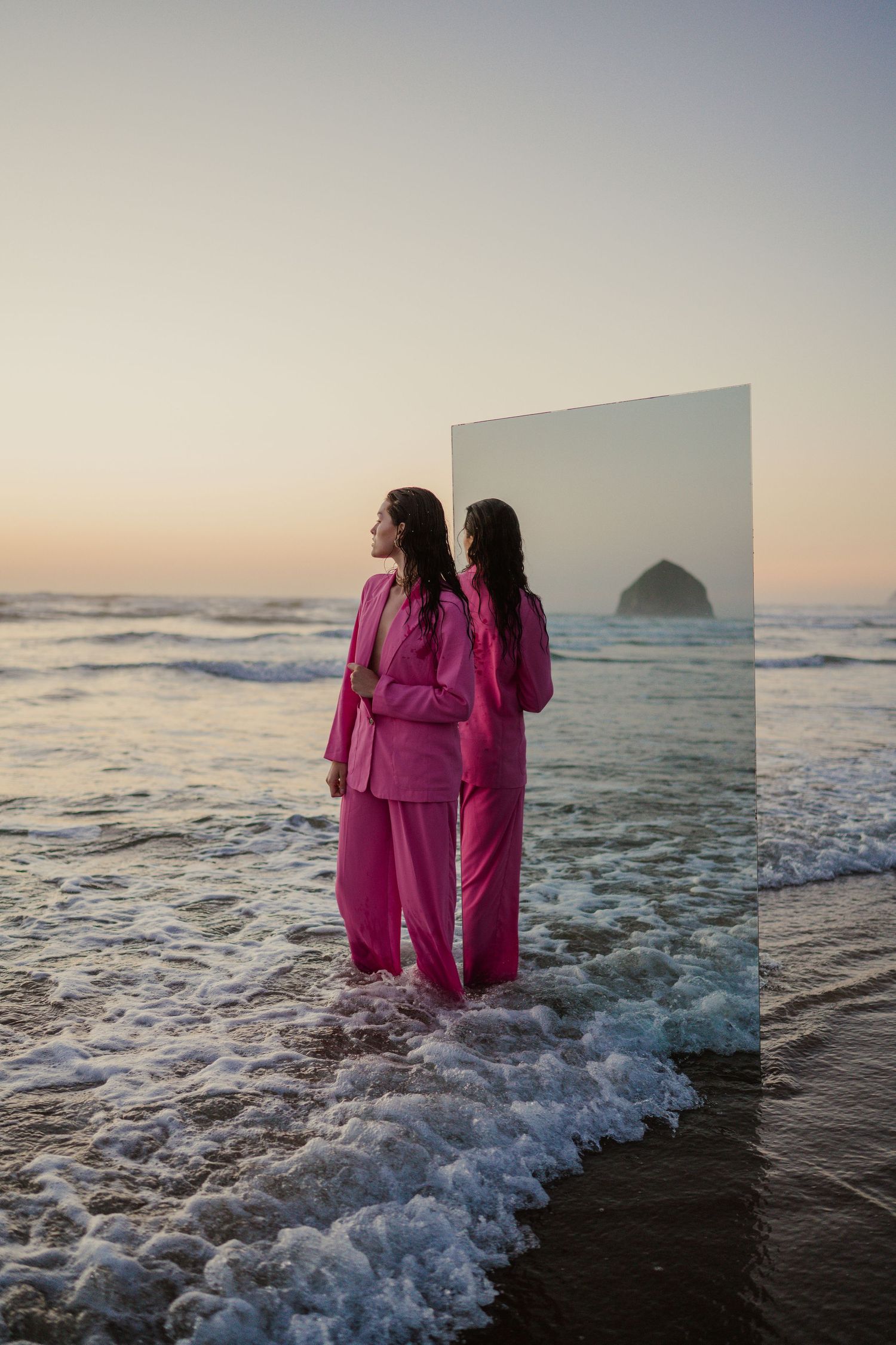 Artistic beach scene with person in pink suit and mirror reflection at sunset with ocean waves and rock formation in background.