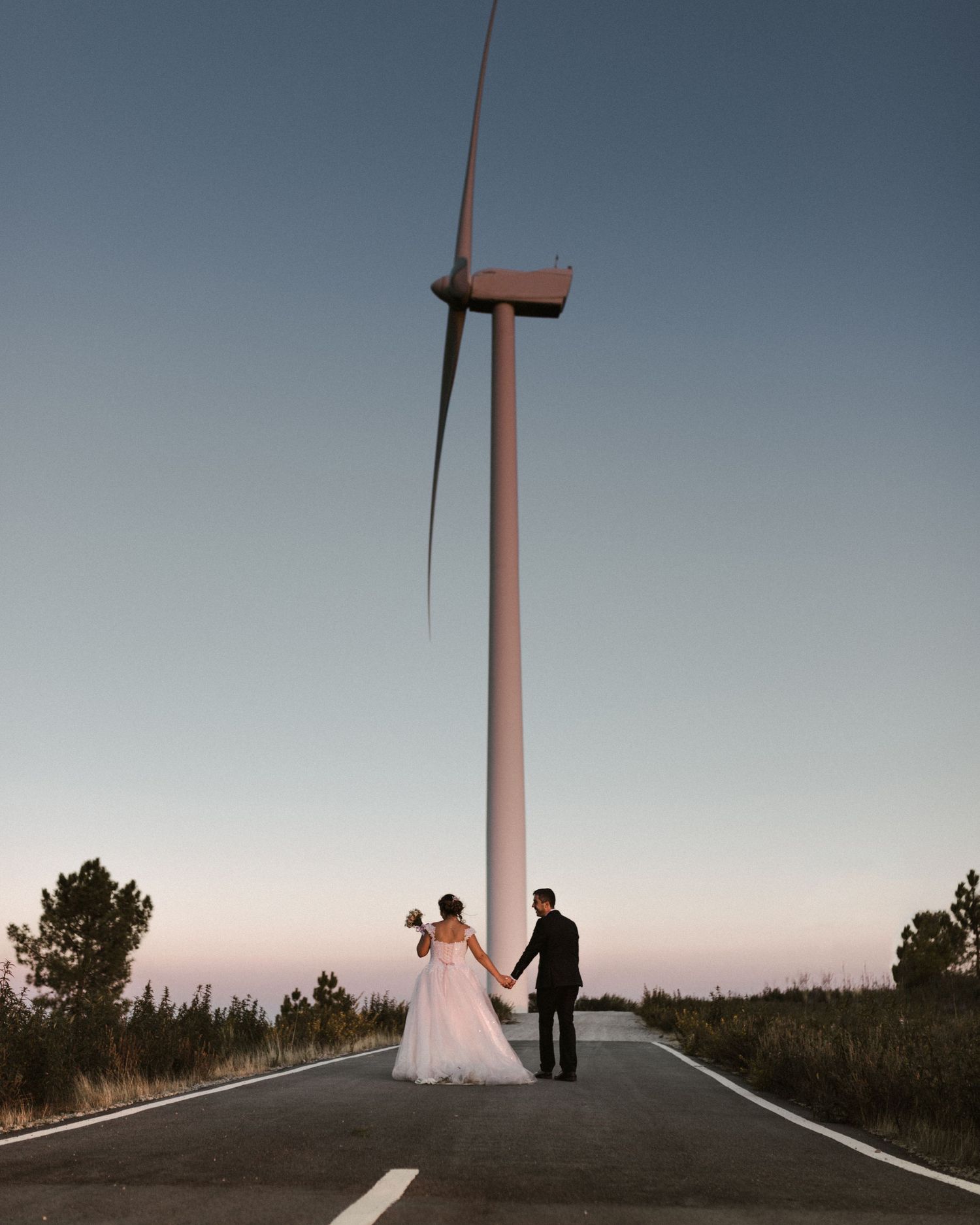 A couple stands on a road with a massive wind turbine rising into the dusky sky.