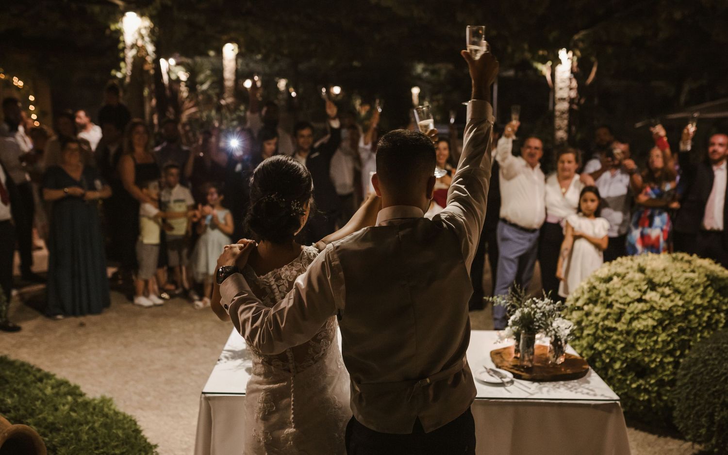 Couple raises sparklers at night during wedding reception as guests cheer with lights in background.