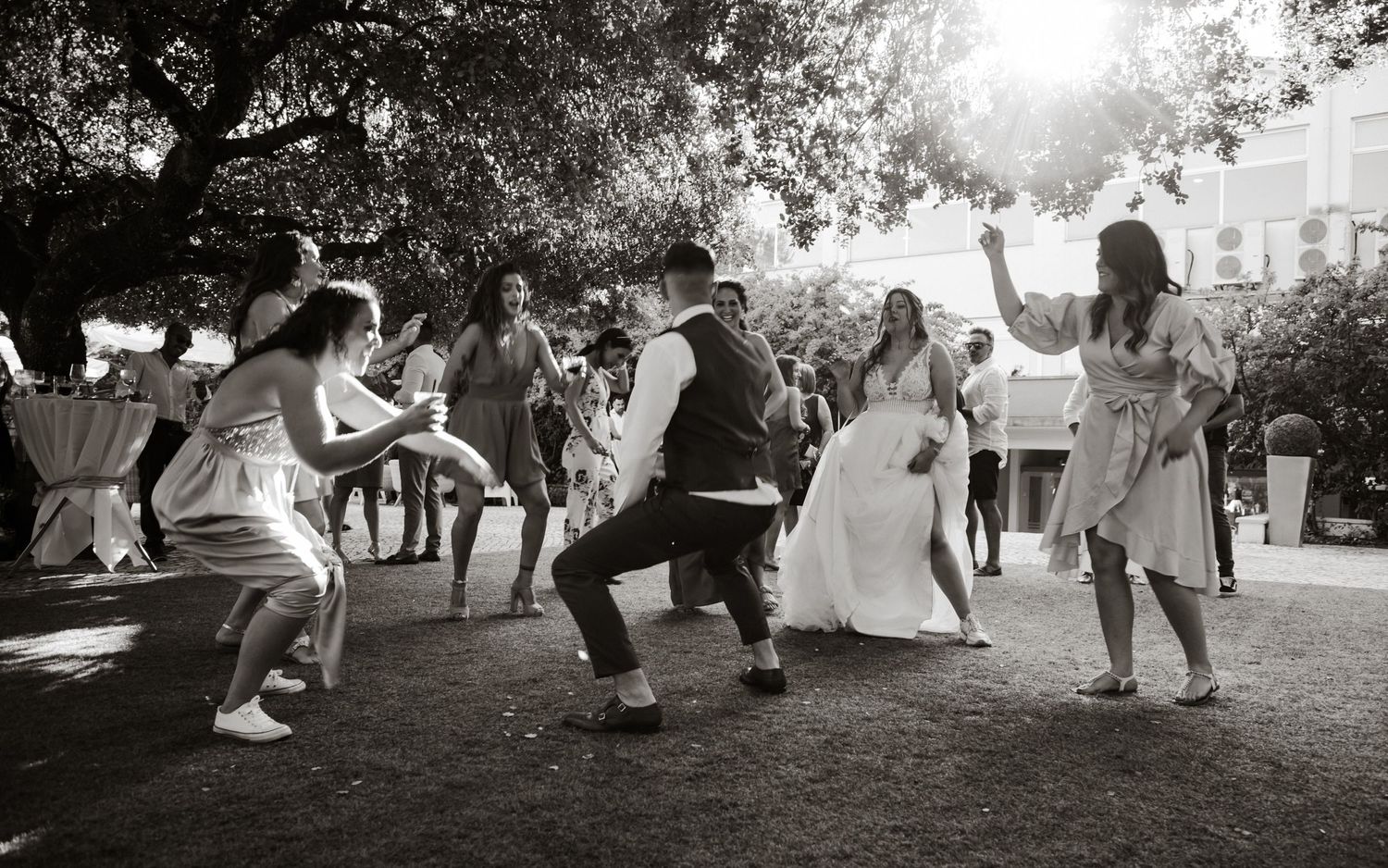 Wedding guests dance joyfully on a lawn during a black and white outdoor celebration.