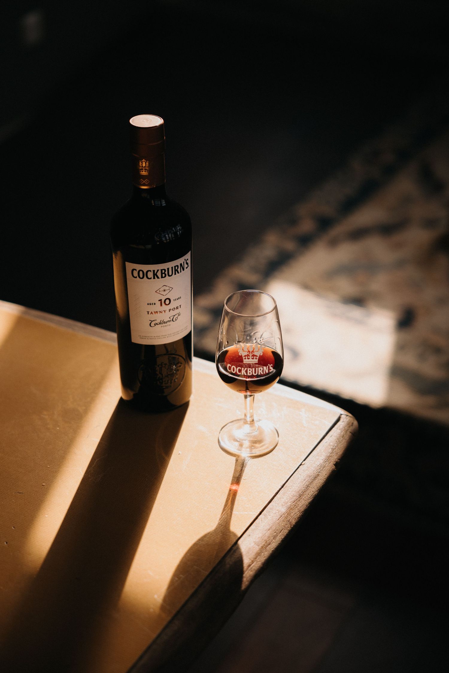 A bottle of Cockburn's port wine and filled glass casting dramatic shadows in moody lighting.