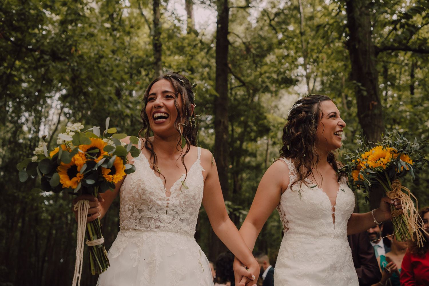 Happy couple in wedding dresses holding sunflower bouquets in a forest celebration.