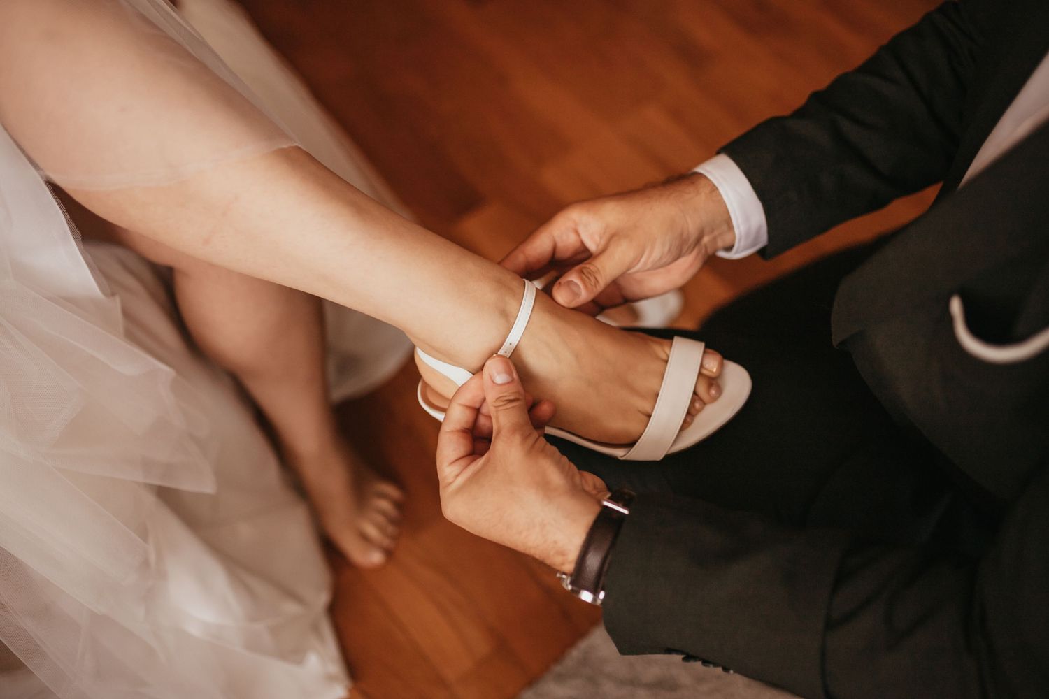 Hands fastening a delicate white wedding shoe buckle on hardwood floor.