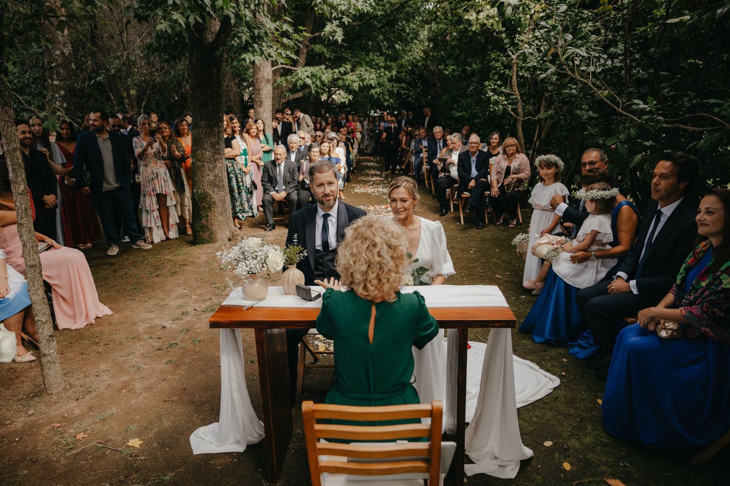 Outdoor wedding ceremony in a wooded setting with guests gathered around a signing table.