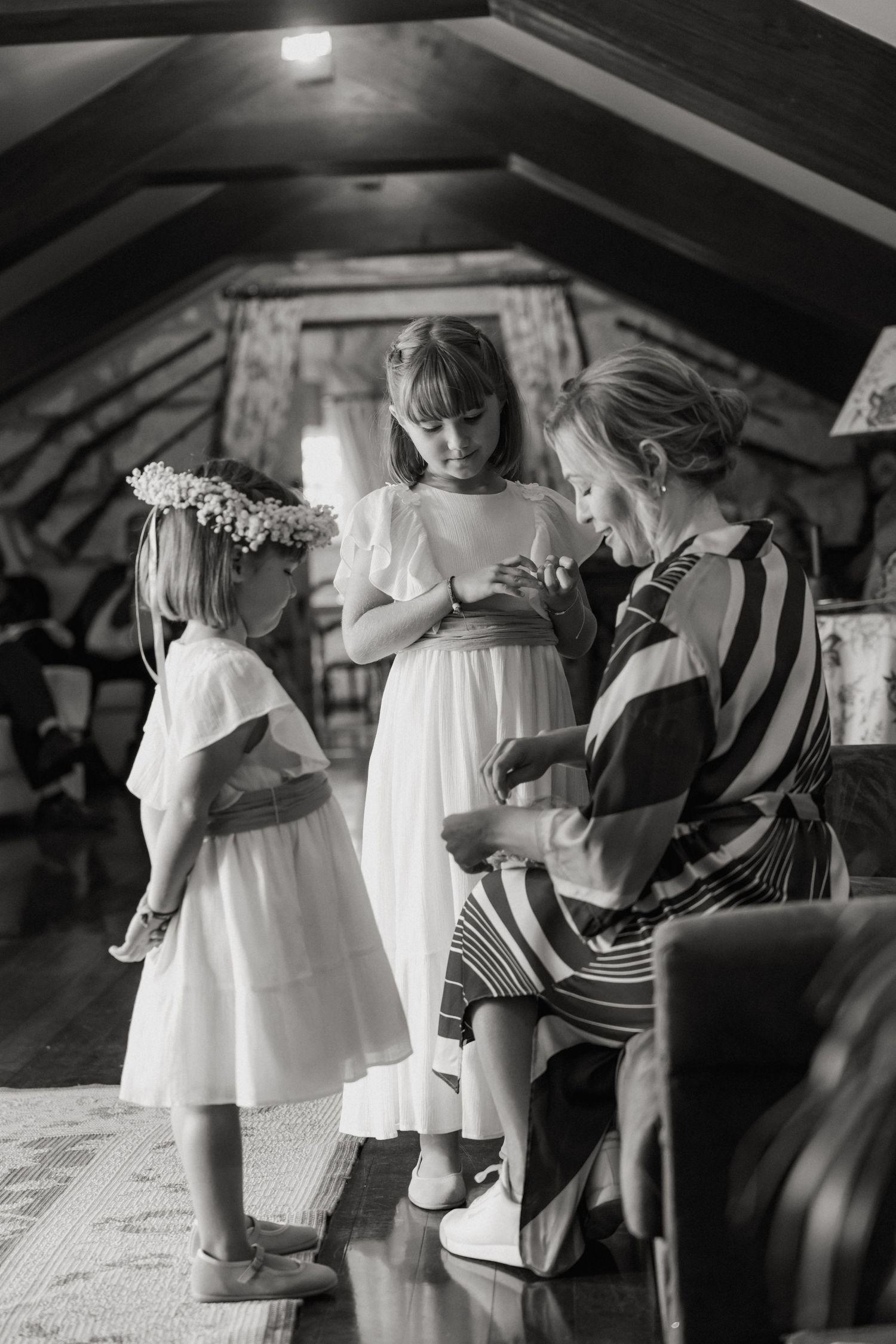 Three people in white dresses gather together during wedding preparations in an attic setting.