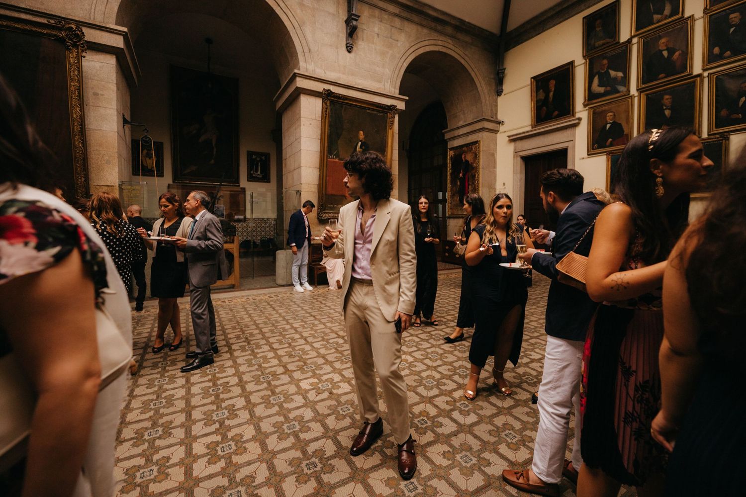 Wedding guests mingling in a historic venue with arched corridors and portrait-lined walls.
