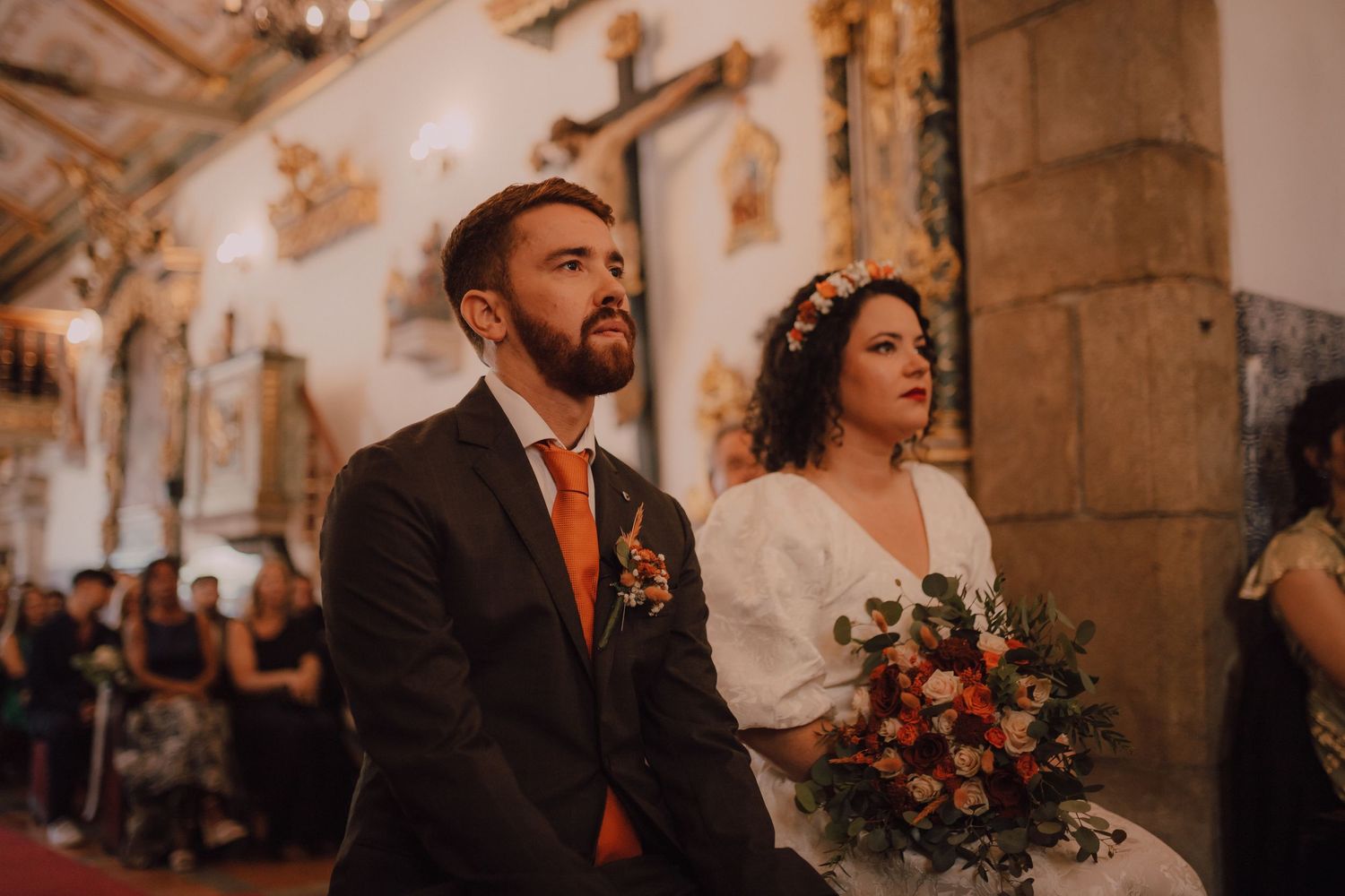 A couple stands at the altar in an ornate church, the bride in white holding a colorful bouquet.
