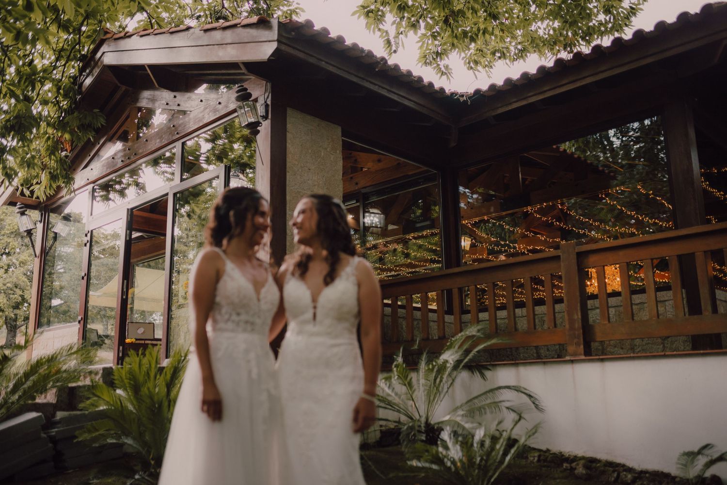 Two people in white wedding dresses share an intimate moment on a covered patio surrounded by lush greenery.