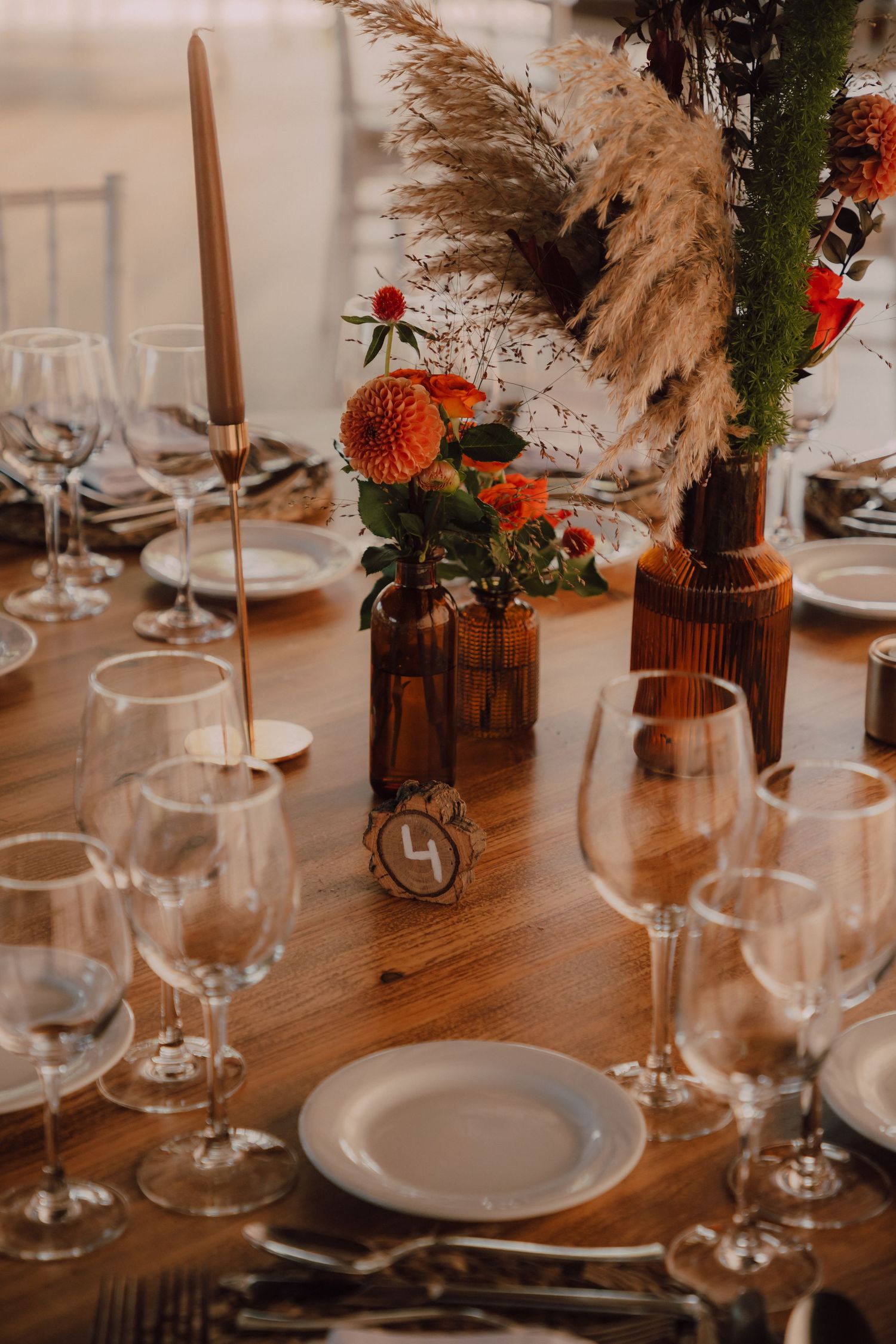 Wedding table decor featuring amber glass vases with orange flowers and pampas grass centerpieces.