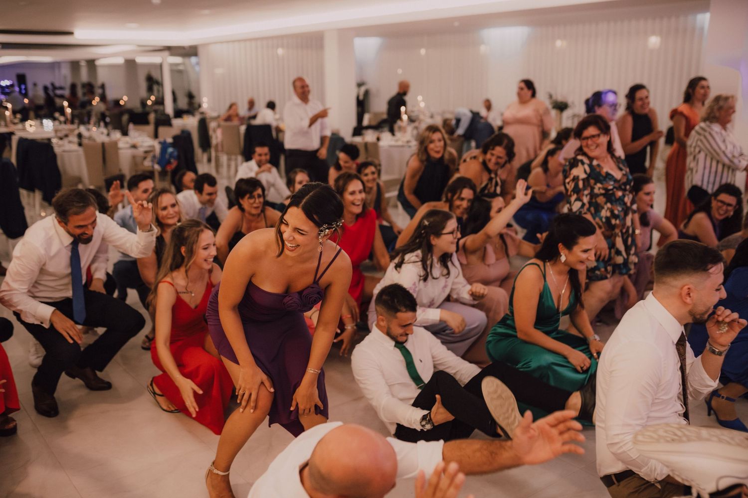 Large group of wedding guests dancing and celebrating at indoor reception.