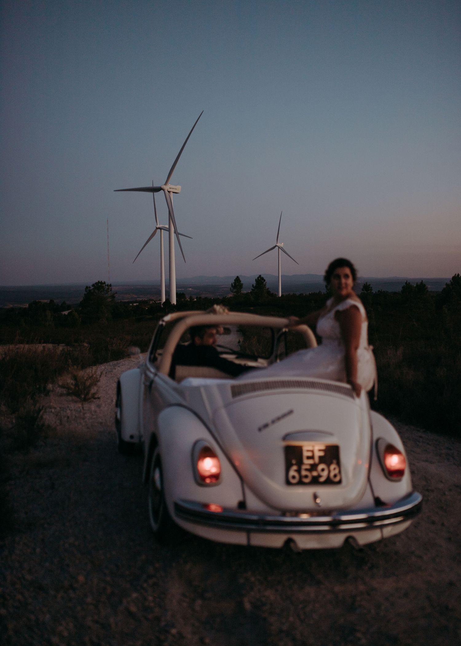 A classic white Volkswagen Beetle parked near wind turbines at sunset.