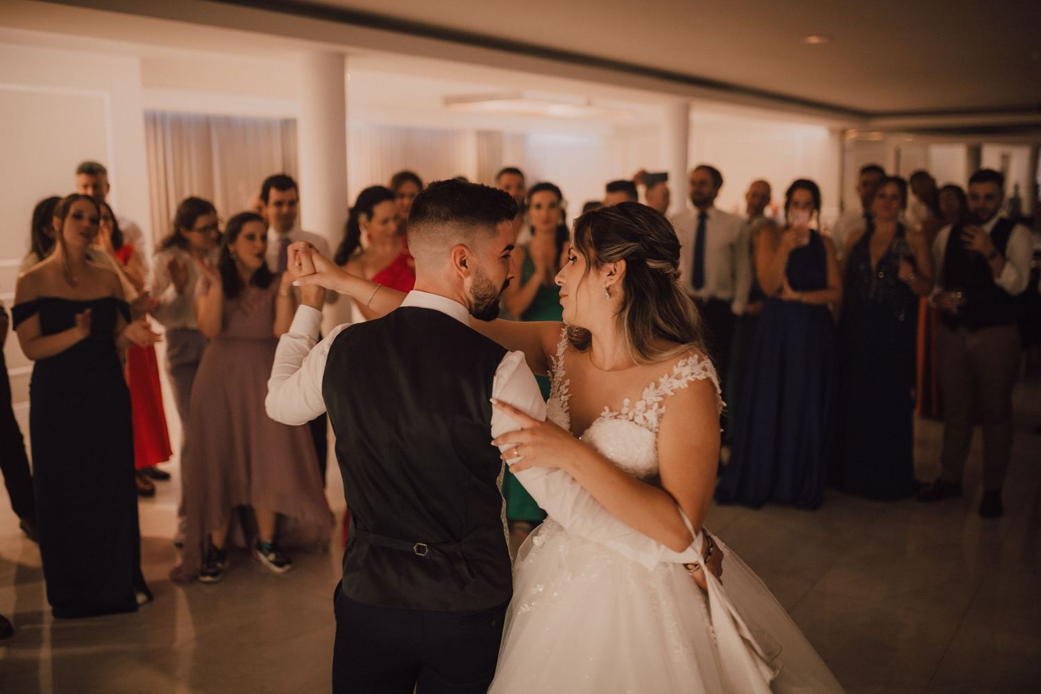 Couple shares their first dance at wedding reception surrounded by guests with cameras.