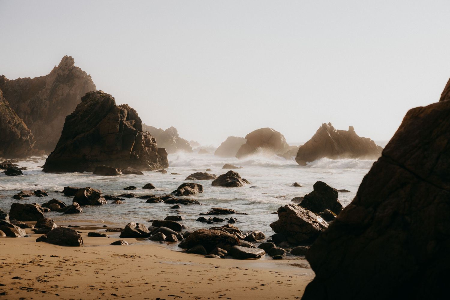 Misty rocky coastline with large boulders scattered across a sandy beach during golden hour.