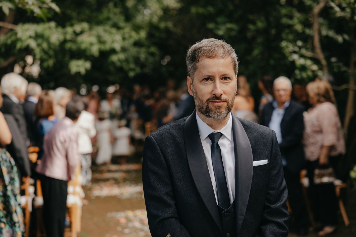A formal wedding guest in a dark suit and tie at an outdoor ceremony.