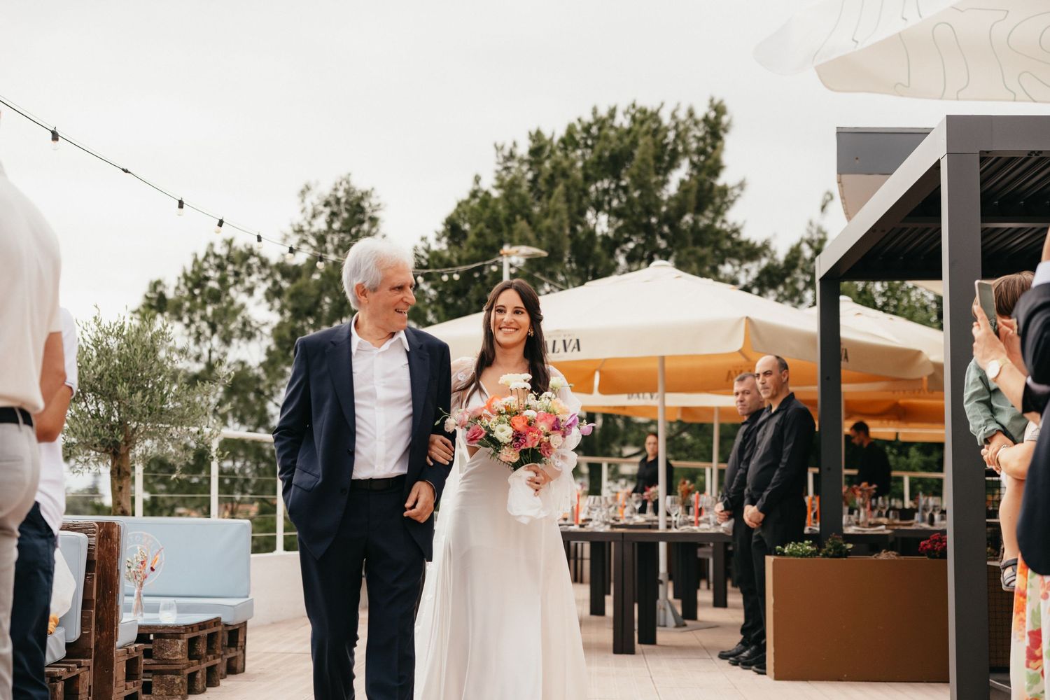 An outdoor wedding processional on a sunny deck with orange umbrellas in the background.