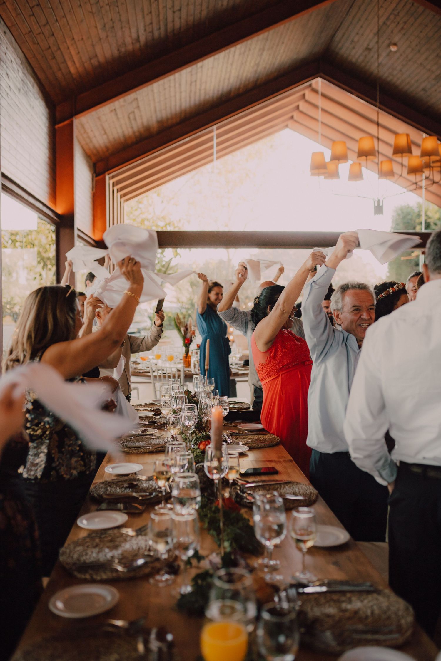 Wedding guests celebrate with raised hands during a dinner reception in a rustic venue with wooden beams.