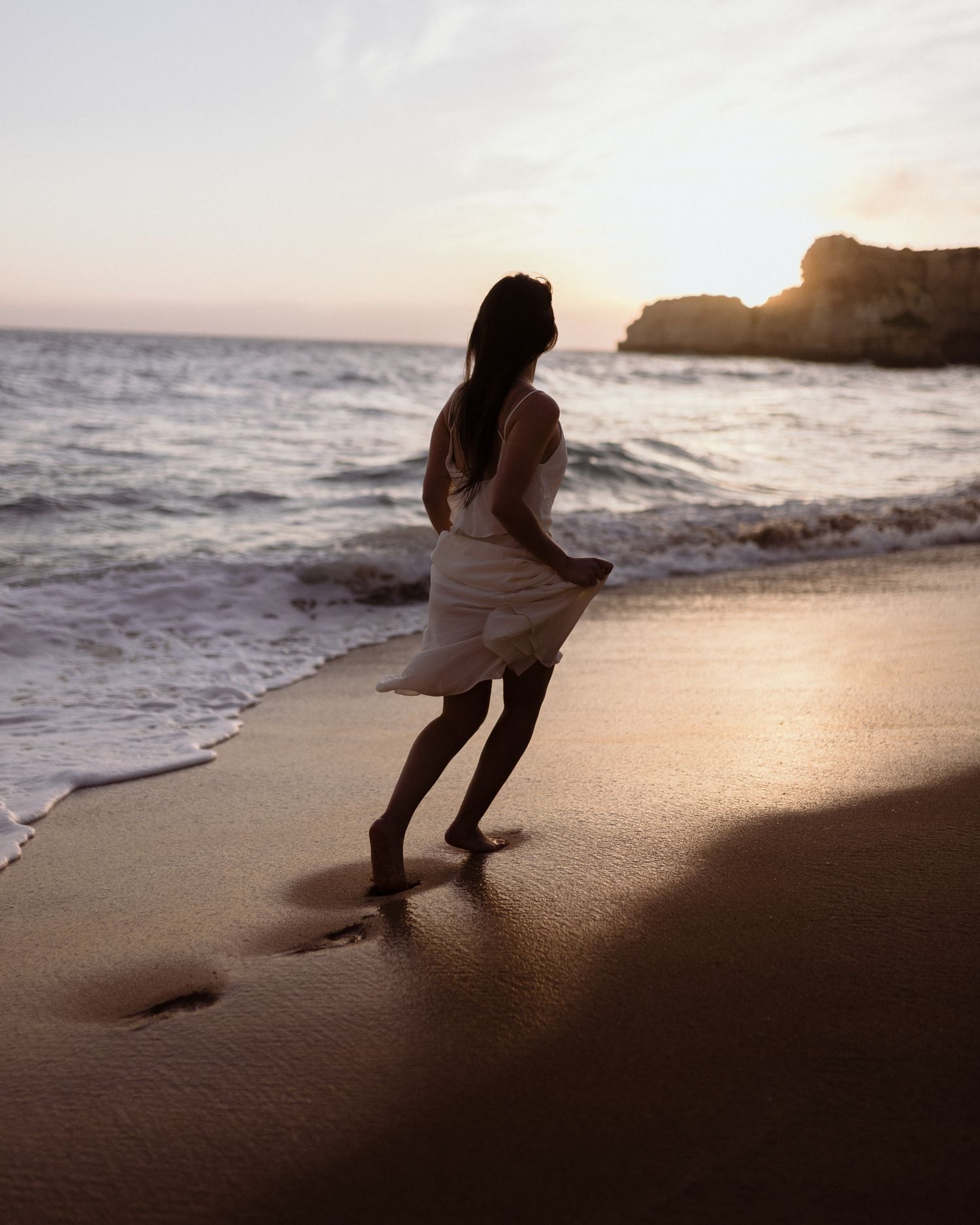 Silhouetted figure dancing on beach at sunset near coastal cliffs.