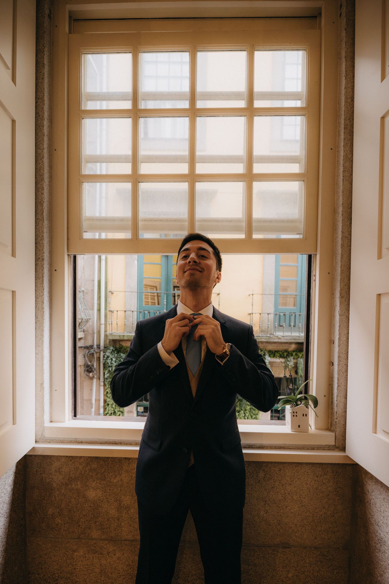 Person in dark suit adjusts tie while standing by large window with natural light.