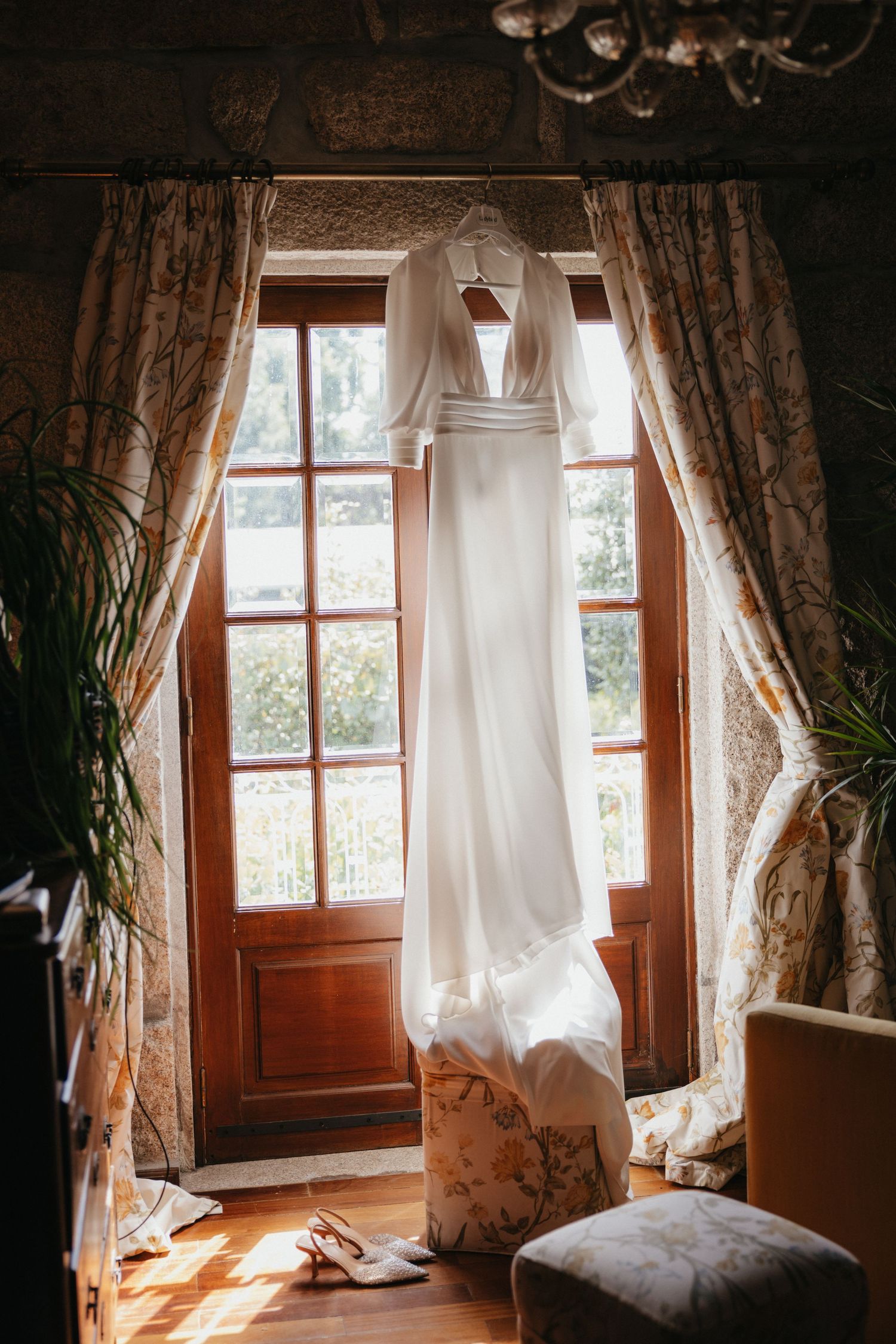 Elegant white wedding dress hanging in front of a sunlit window with draped curtains.