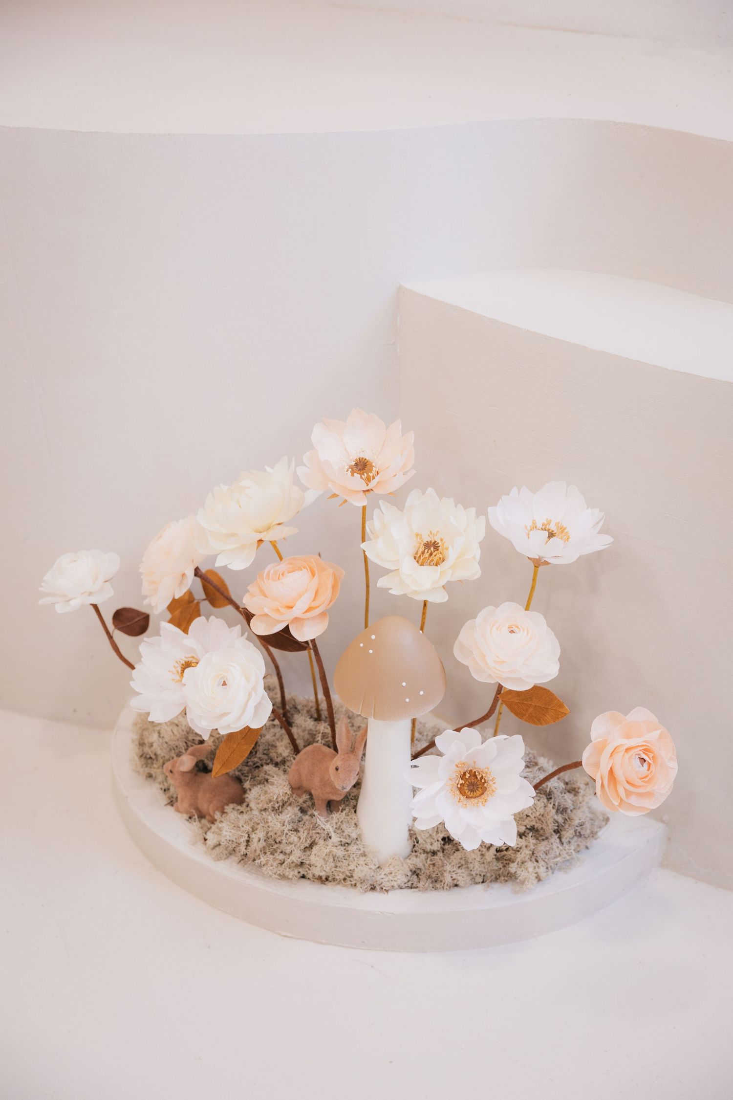 A series of soft pink and peach flowers arranged in a white ceramic bowl with dried foliage and sand.