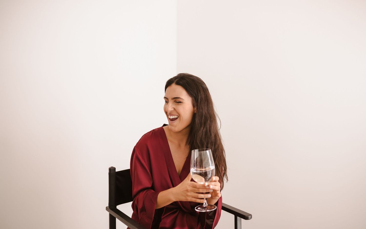 Person laughing while holding wine glass against minimal white backdrop.
