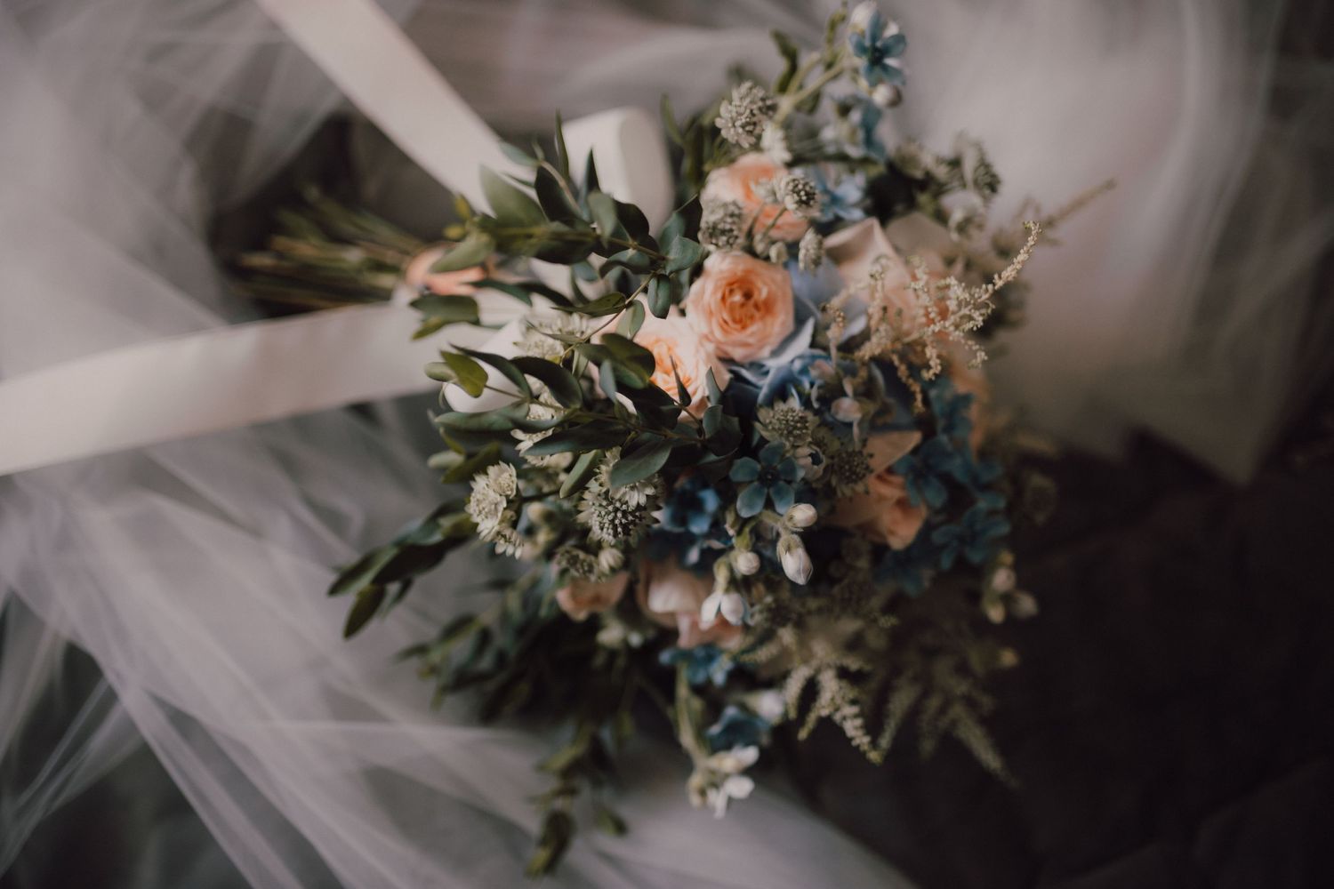 Delicate wedding bouquet with peach roses and blue florals against white tulle.