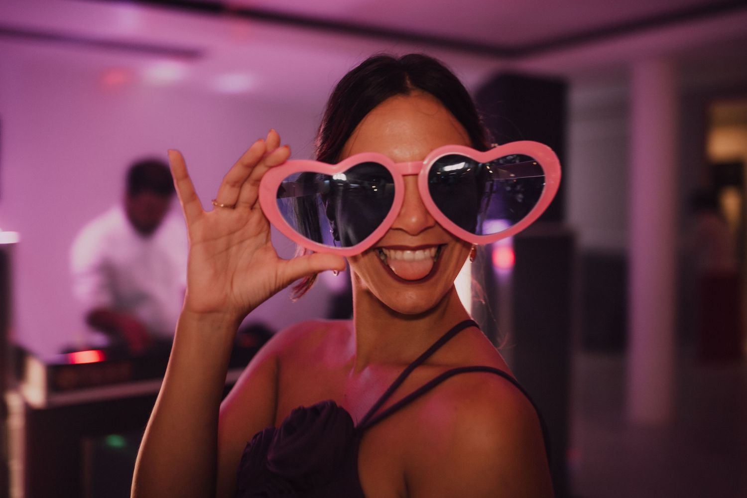 A festive wedding guest poses playfully with heart-shaped sunglasses in pink party lighting.