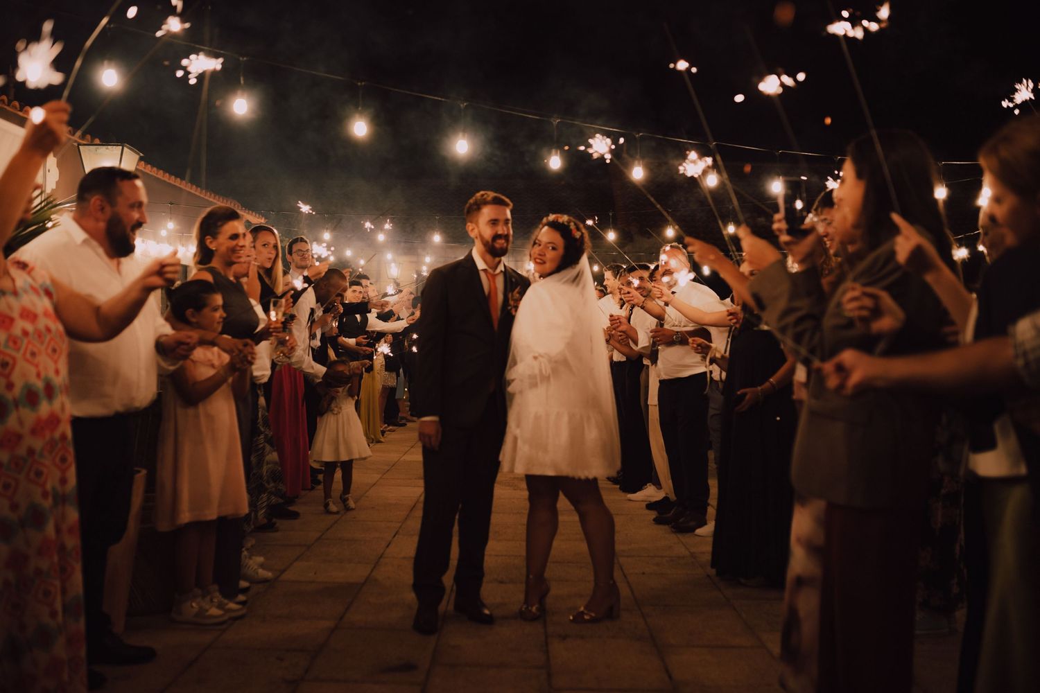 Guests hold sparklers creating a magical tunnel at a nighttime wedding celebration.
