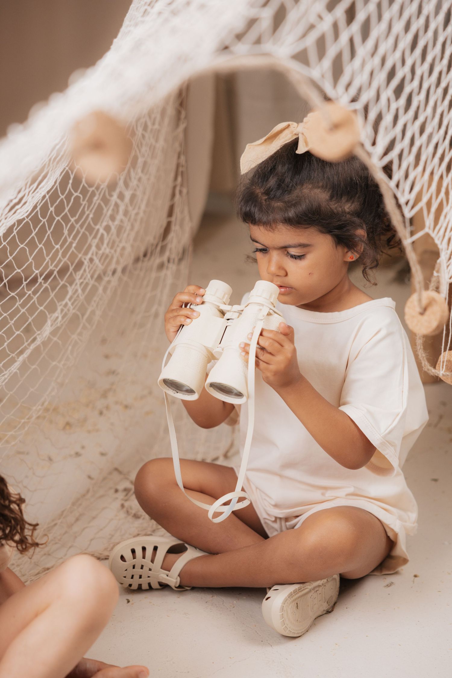 Small child in white outfit plays on floor near hammock netting in warm neutral tones.