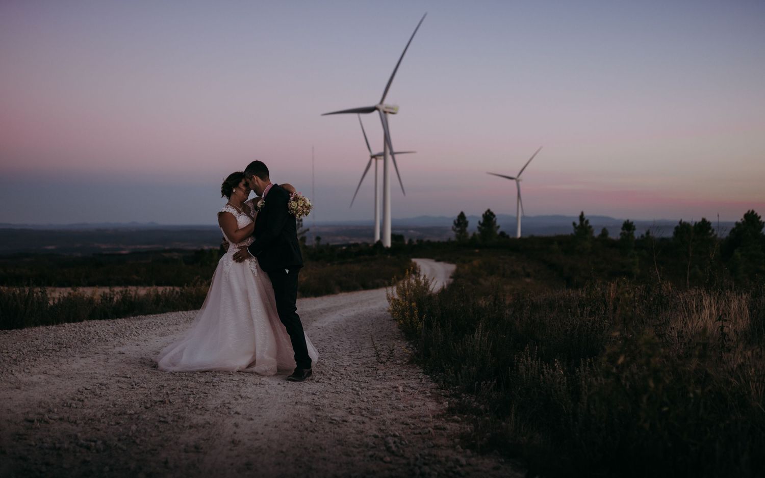 Romantic wedding couple standing on dirt road with wind turbines at sunset.