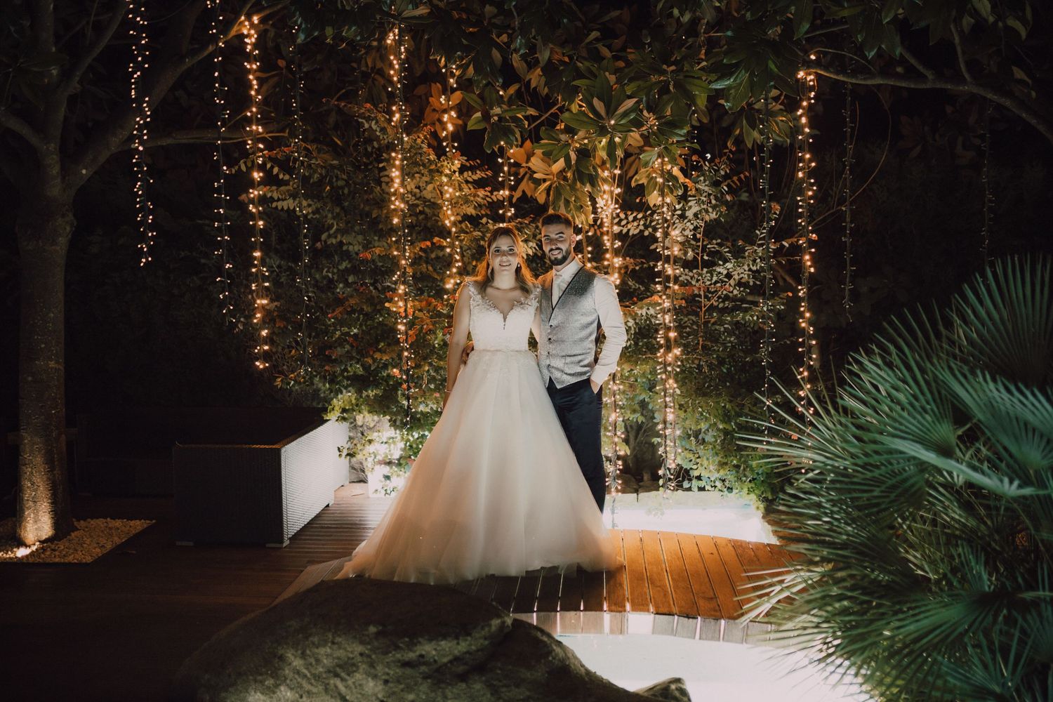 Couple poses under romantic fairy light display at evening wedding venue.