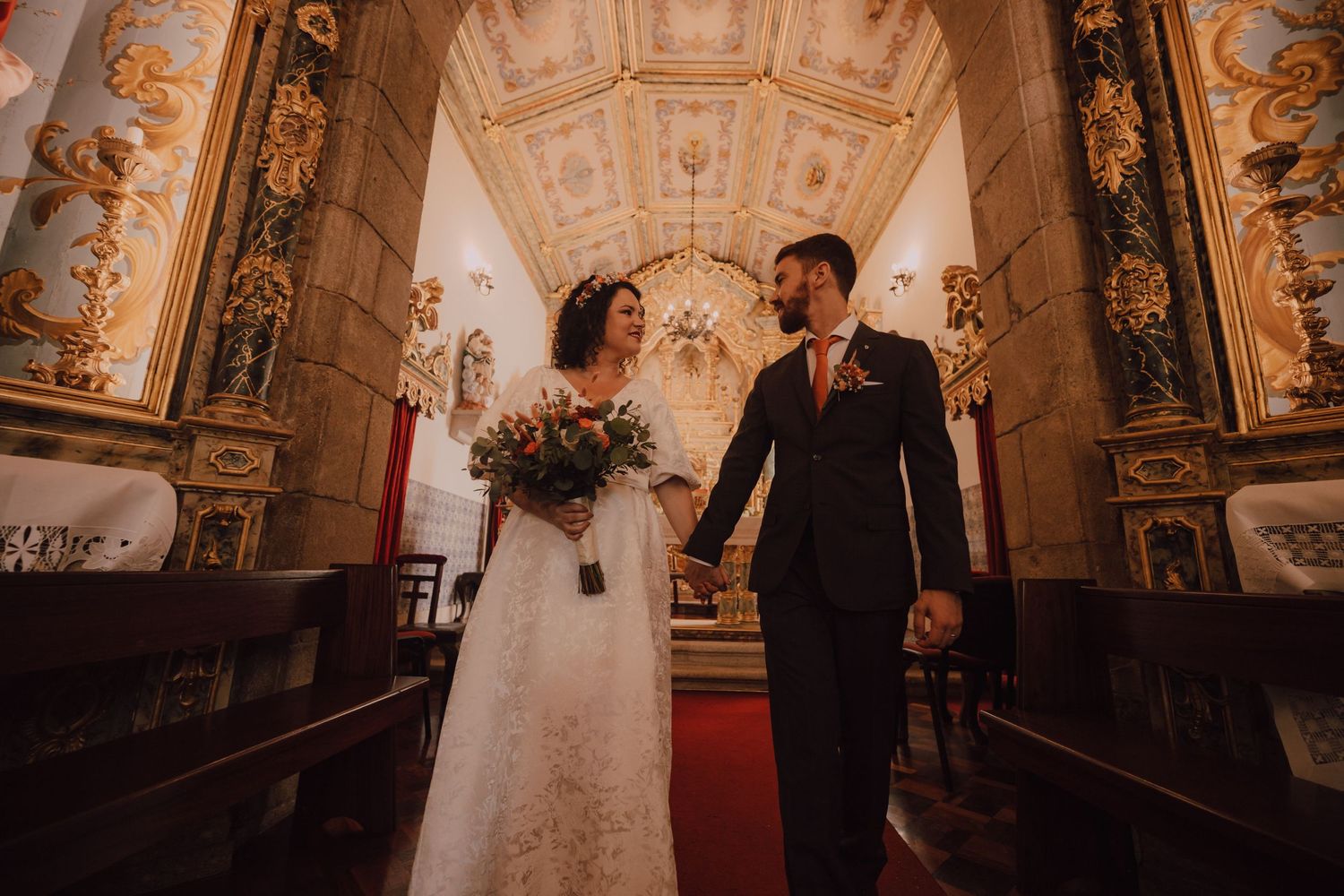 A couple walks down ornate church aisle with gilded details and red carpet.
