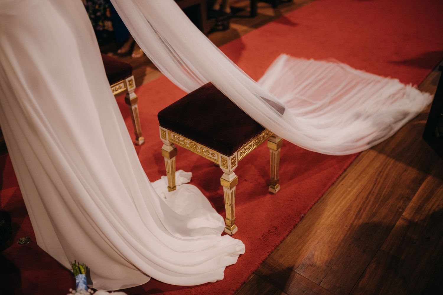 White wedding dress train flowing over ornate gold footstool on red carpet in church.