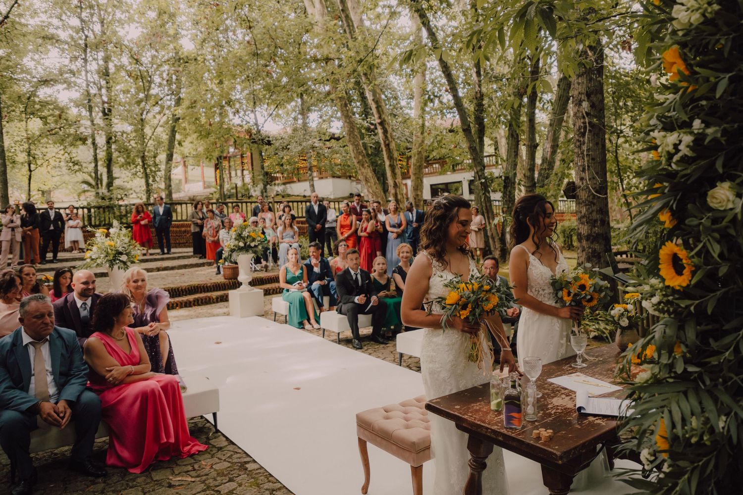 Outdoor wedding ceremony setup with wooden chairs, sunflower decorations, and guests in colorful attire.
