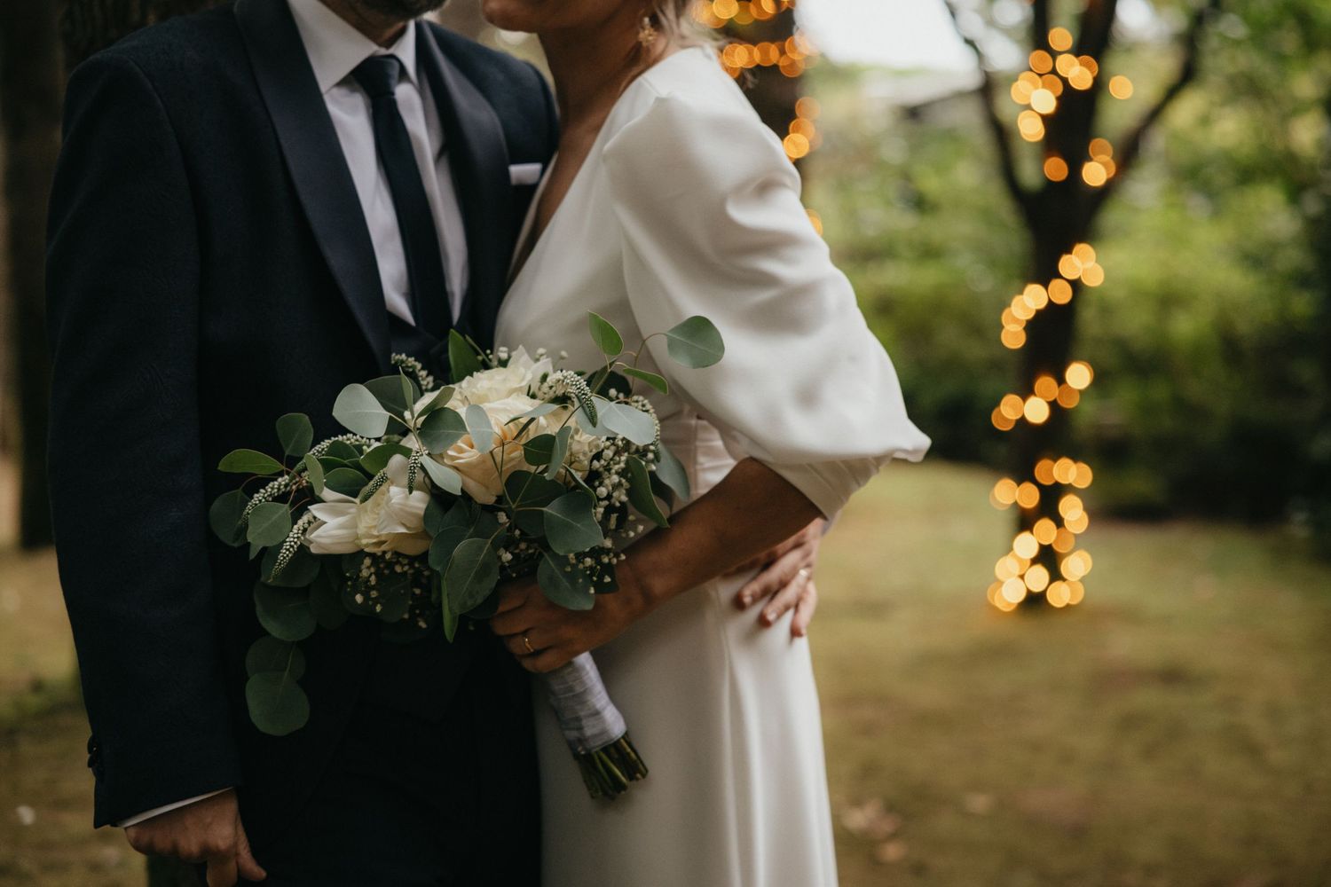 Elegant wedding photo with white bouquet and twinkle lights in garden setting.