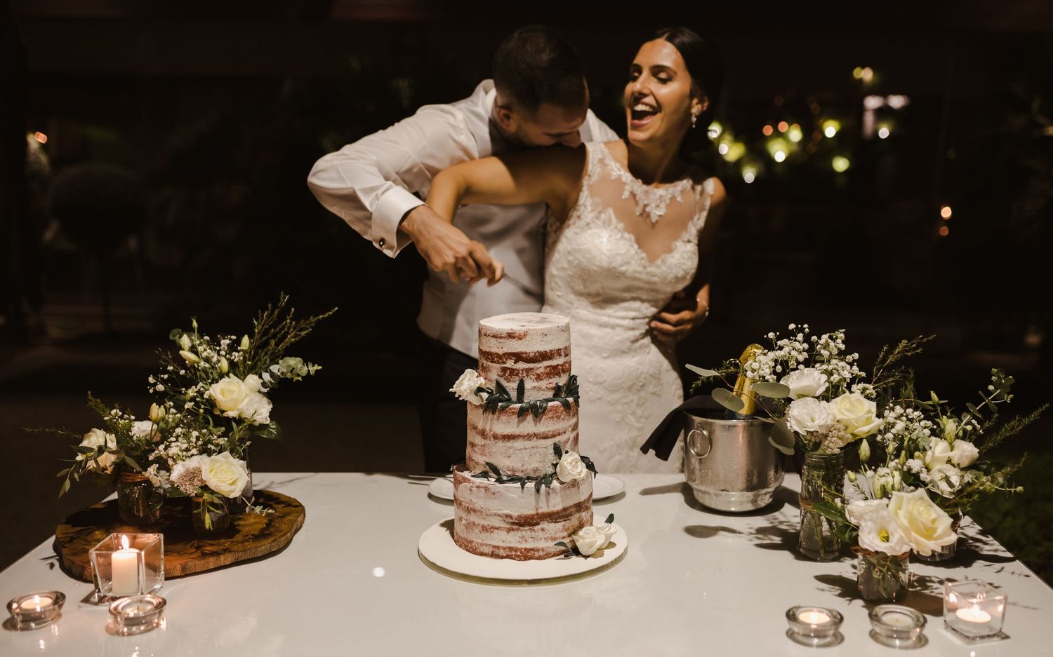 Newlyweds cut into their elegant wedding cake decorated with white flowers and greenery.