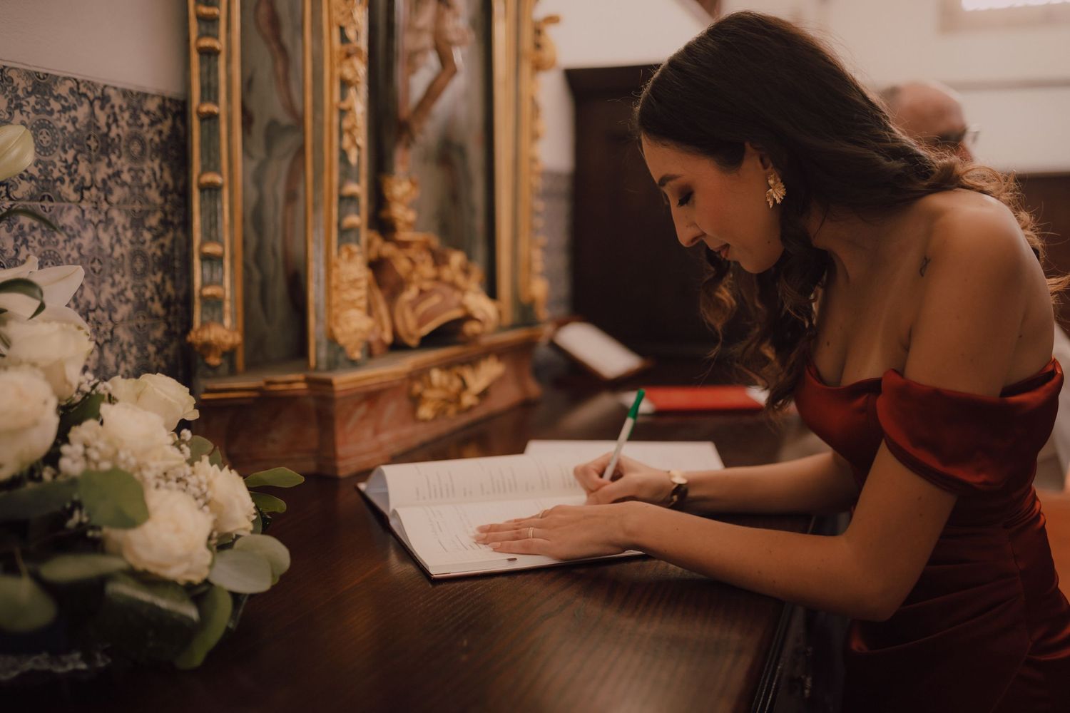 Someone in burgundy dress writing in guestbook at elegant vintage desk.