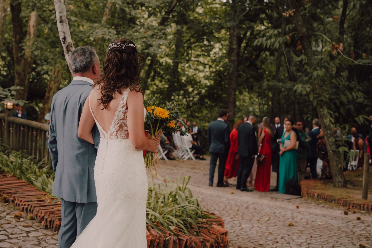 Wedding guests gather on a pathway surrounded by trees and natural landscaping.