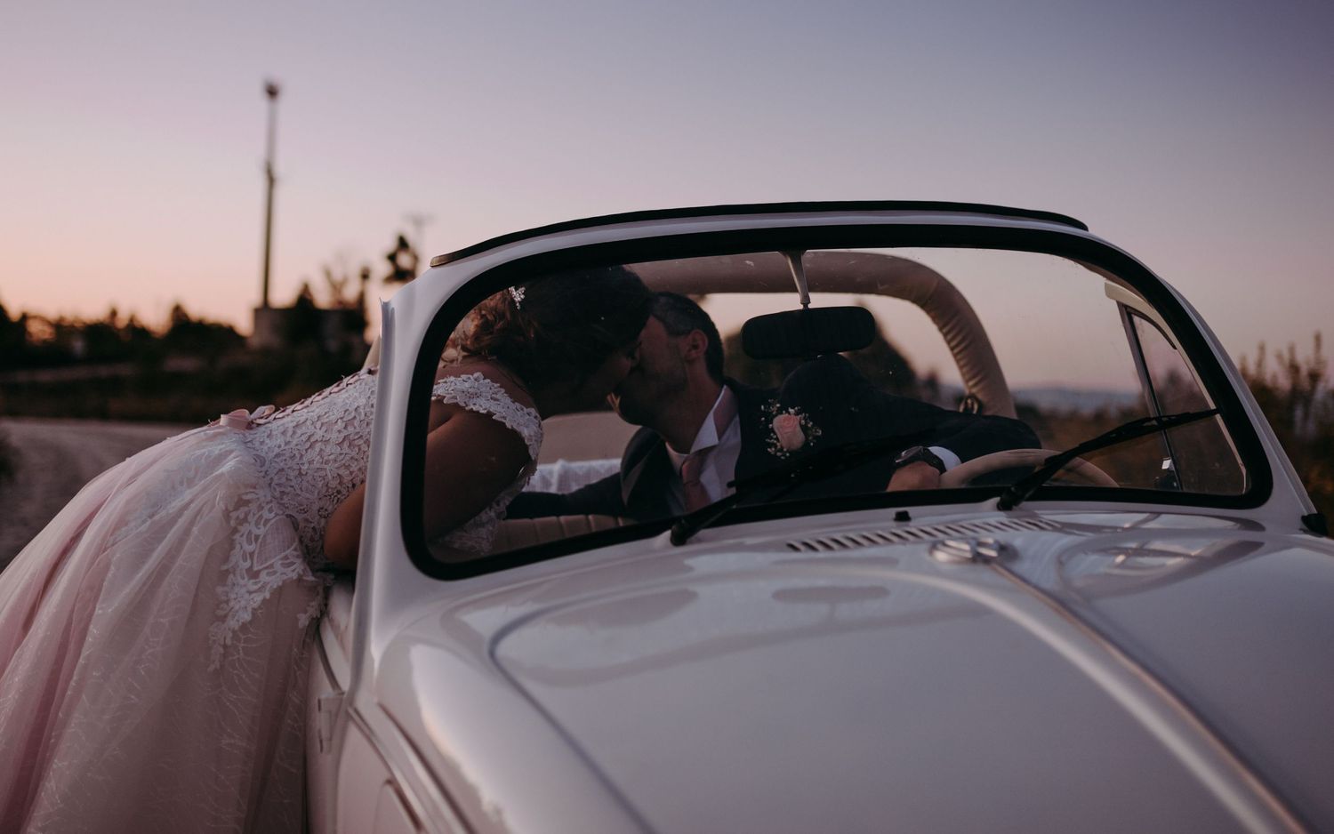 Classic white Volkswagen Beetle with wedding decorations against sunset sky.