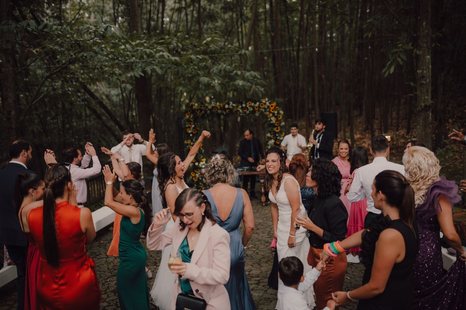 Wedding guests dance and celebrate on an outdoor dance floor surrounded by trees and string lights.