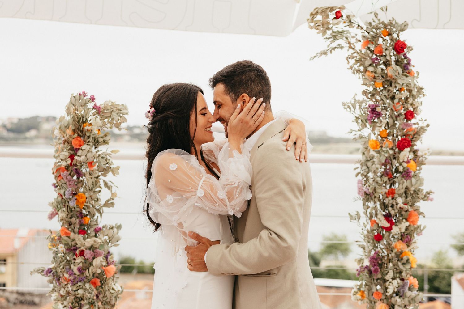 Romantic wedding moment under floral arch with ocean backdrop.